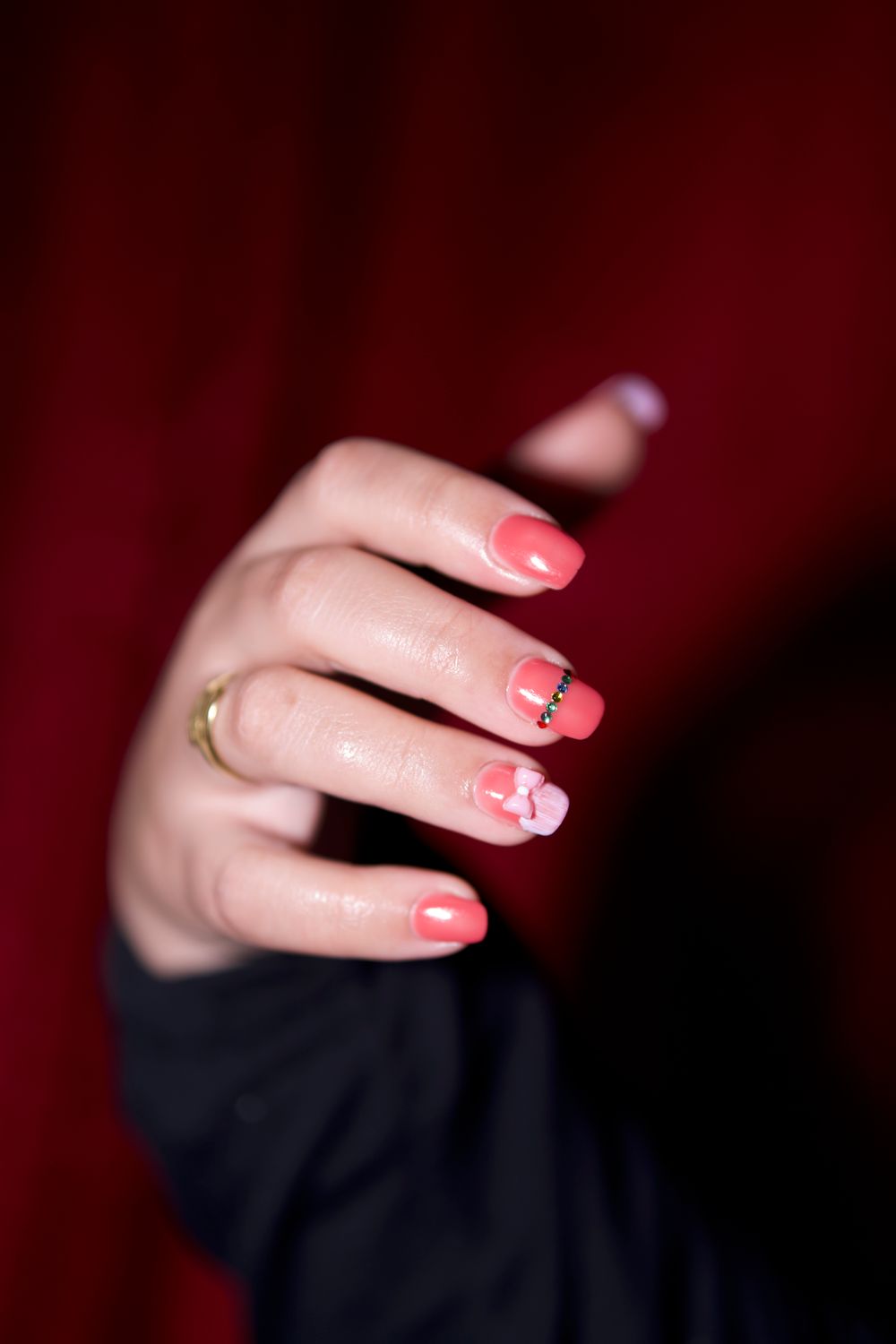 Hand met koraalroze nagels met een strikje en fijne lijntjes, tegen een donkerrode achtergrond