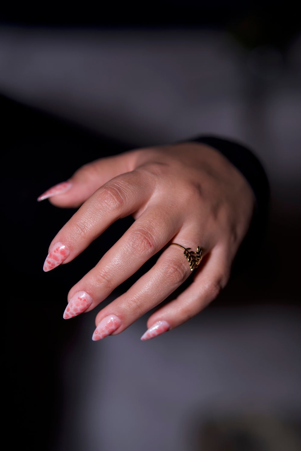 Hand met amandelvormige roze nagels met een fijn patroon en een ring, tegen een donkere achtergrond